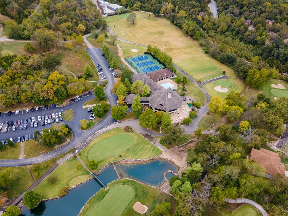 This wide-angle exterior frames the golf course, tennis courts, main building, parking area, and a pond. The lush greenery and open layout evoke a sense of space and recreational abundance.