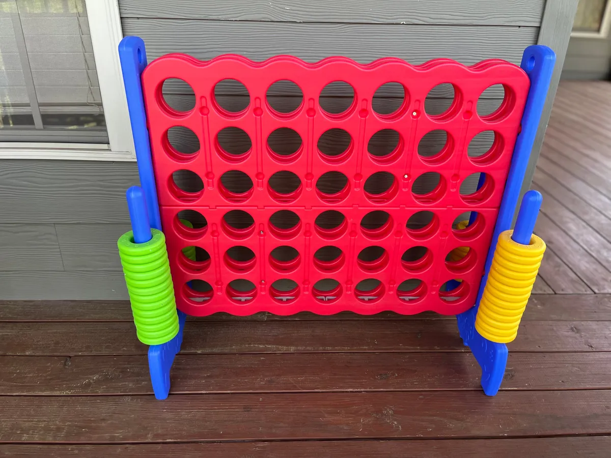A bright, joyful scene featuring a life-size Connect 4 game set up on a wooden deck floor, with vibrant colors and an inviting atmosphere perfect for laughter-filled evenings and family fun.