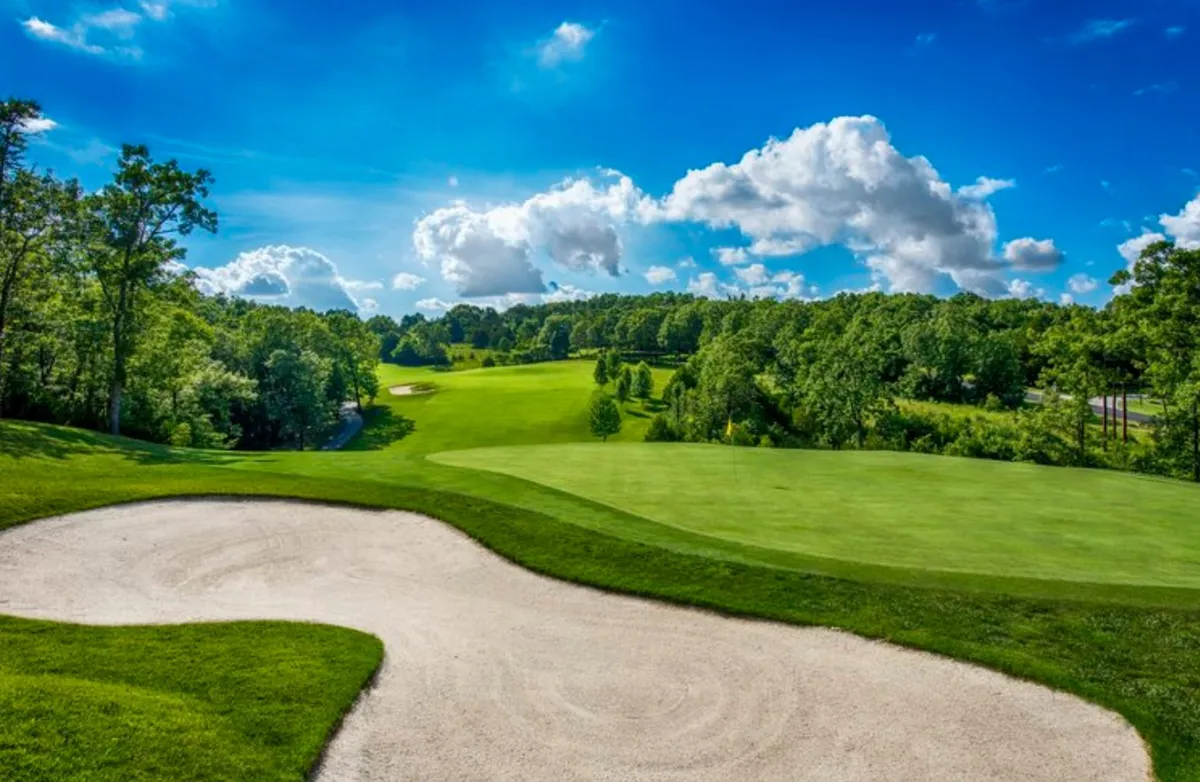 A wide, serene view of the golf course with lush green grass, a prominent sand bunker, rolling hills, and a blue sky dotted with clouds. The scene radiates tranquility and the promise of a championship golf experience in a beautiful natural setting.