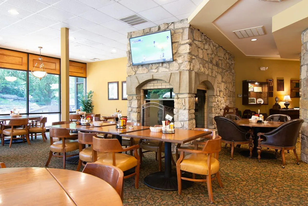 The cozy interior of the clubhouse or grille, featuring a stone fireplace, dining tables, and large windows that let in natural light. The space feels inviting and relaxed, perfect for post-round meals or socializing with friends after a day on the course.