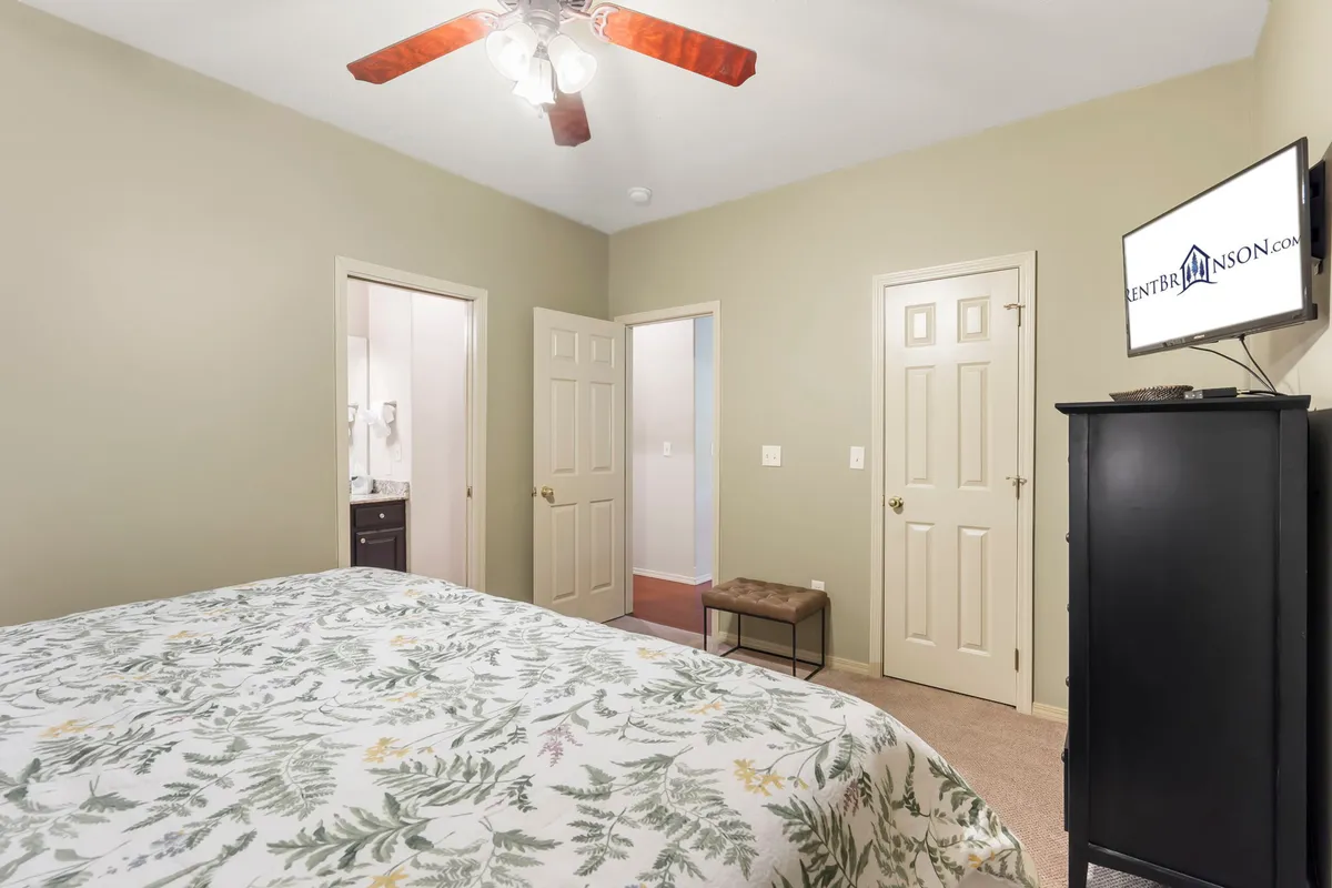 A spacious bedroom with floral bedding on a king-sized bed, ceiling fan, wall-mounted television, and double entry doors. The scene is warm and inviting, ideal for restful getaways or cozy evenings spent inside. The visible TV and plush bedding reinforce the idea of a private haven.