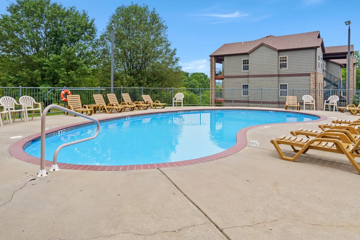 This image shows a sparkling outdoor swimming pool surrounded by lounge chairs and lush green trees. The pool area is adjacent to a building structure, evoking a classic resort atmosphere perfect for families to relax and play. The inviting environment suggests opportunities for swimming, sunbathing, and poolside gatherings.